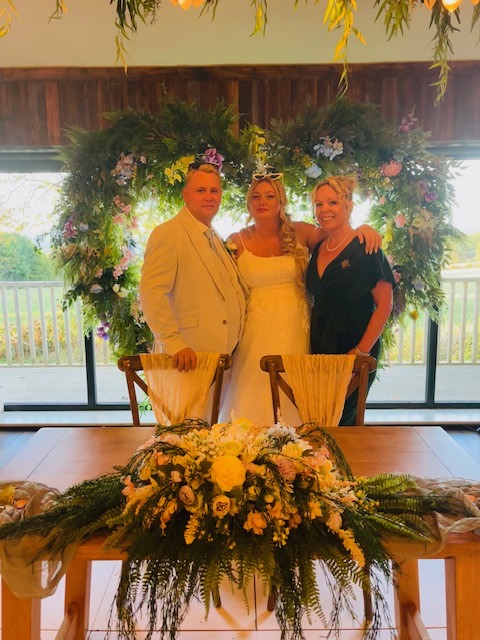 Clare and a wedding couple standing beneath a floral arch behind the ceremony table.