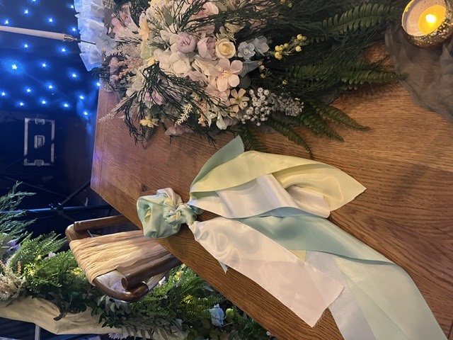 Pale green and ivory handfasting ribbons beside flowers on a wooden ceremony table.
