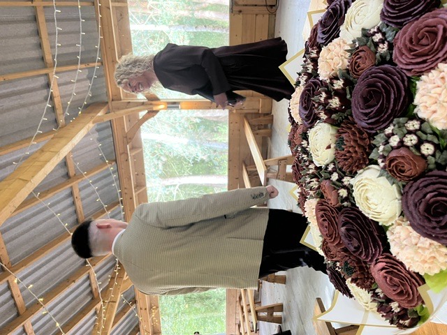 Clare smiling with the groom in a wooden ceremony shelter, with flowers in the foreground.